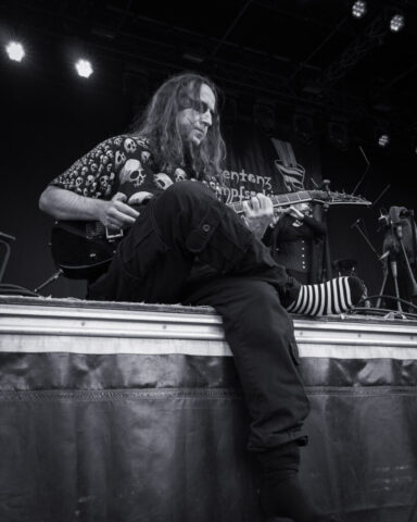 Der Irre sitzt am Bühnenrand und spielt Gitarre. Das Bild ist schwarz weiß.